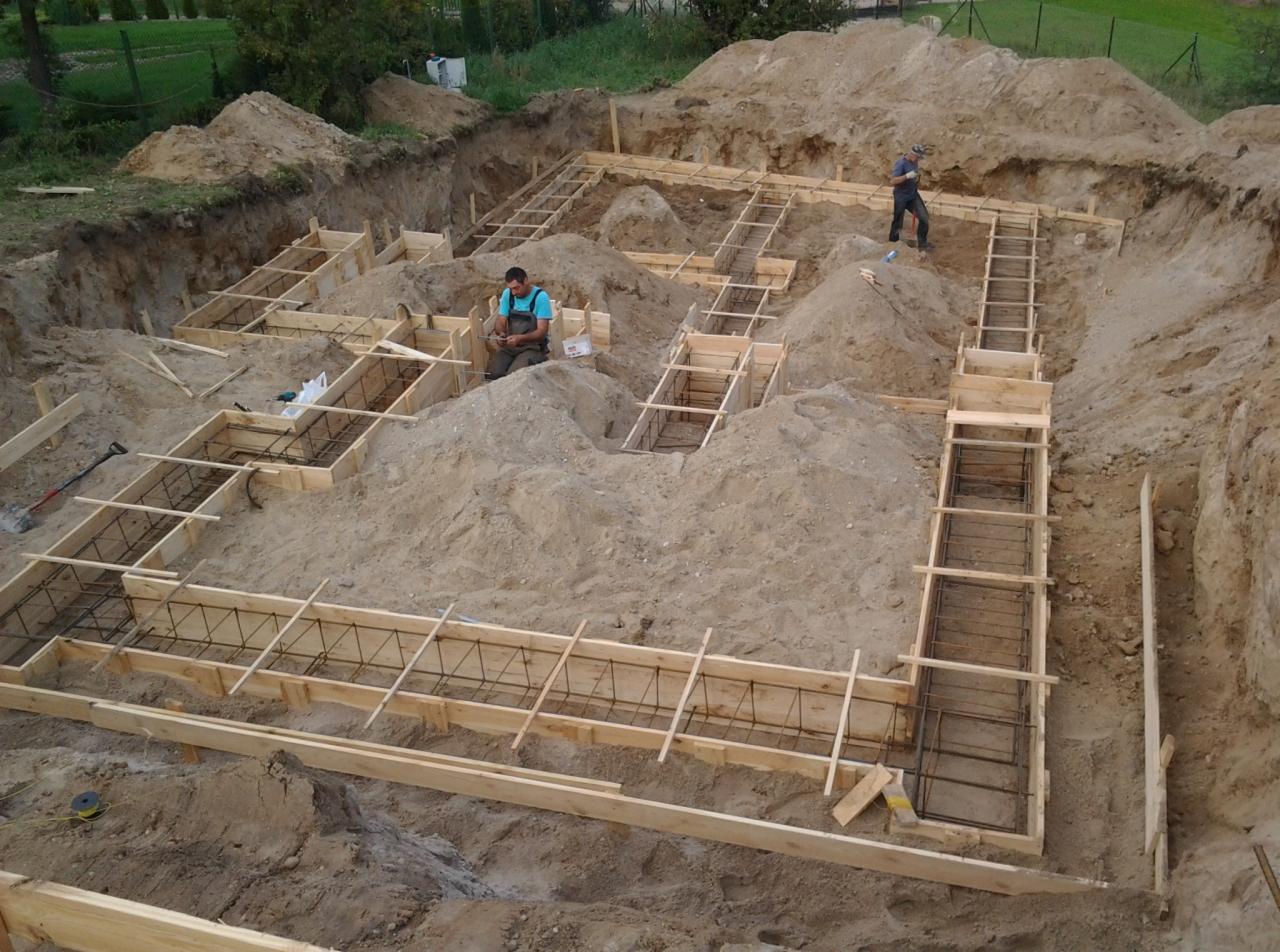 Wykonanie szalunku fundamentów pod budynek w wykopie, widoczni dwaj pracownicy budowlani, jeden siedzi w wykopie przy szalunku, drugi stoi na górze. Szalunek zbrojony stalą. Duże ilości piasku...