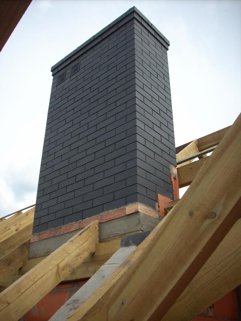 Wysoki komin obłożony grafitową klinkierową cegłą, widoczny na tle konstrukcji dachu w trakcie budowy, z ceglaną bazą i betonowym wykończeniem.