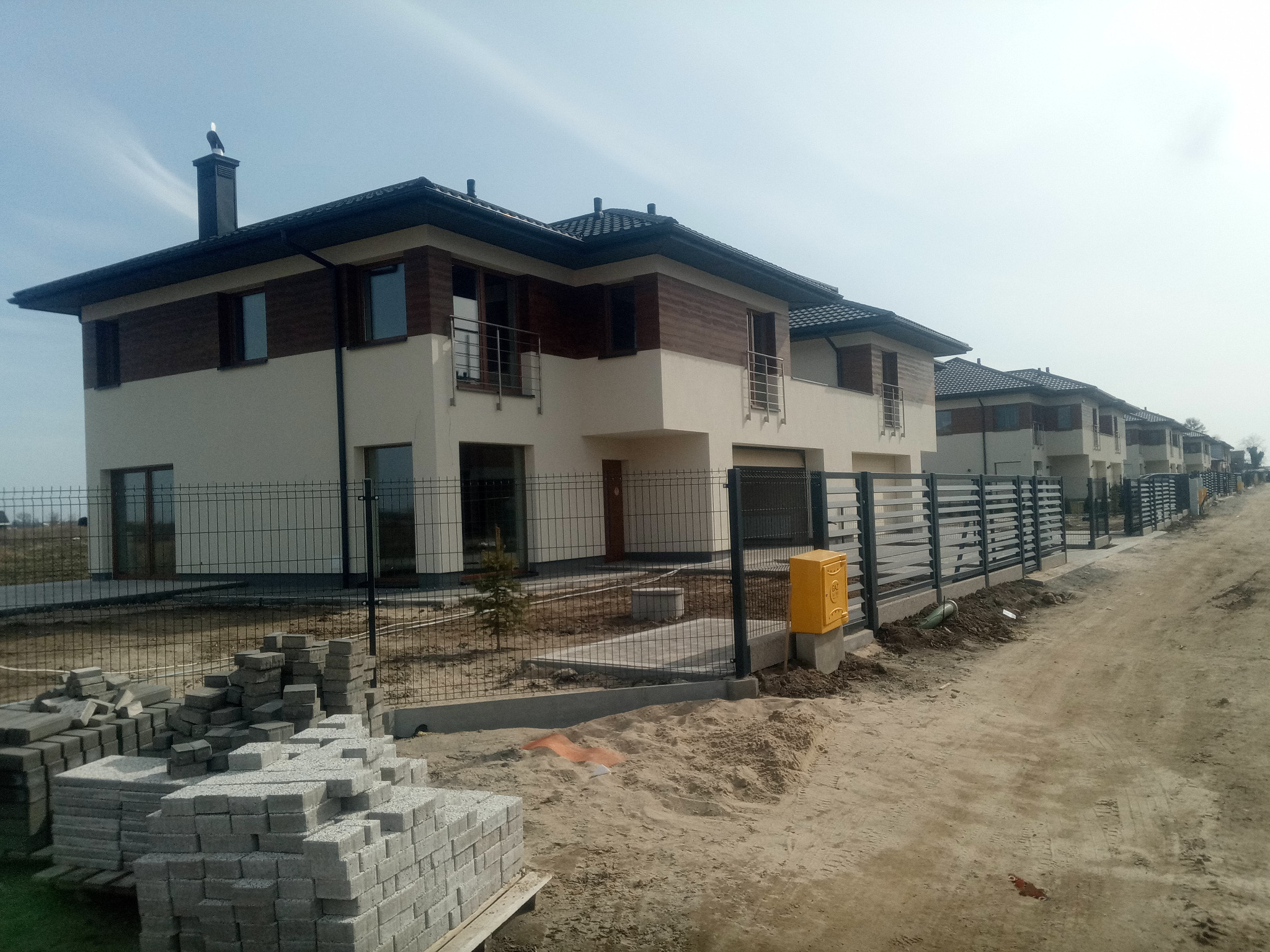 Szereg dwukondygnacyjnych domów w trakcie budowy, z widocznymi stertami kostki brukowej na pierwszym planie i bocianem na kominie jednego z budynków.