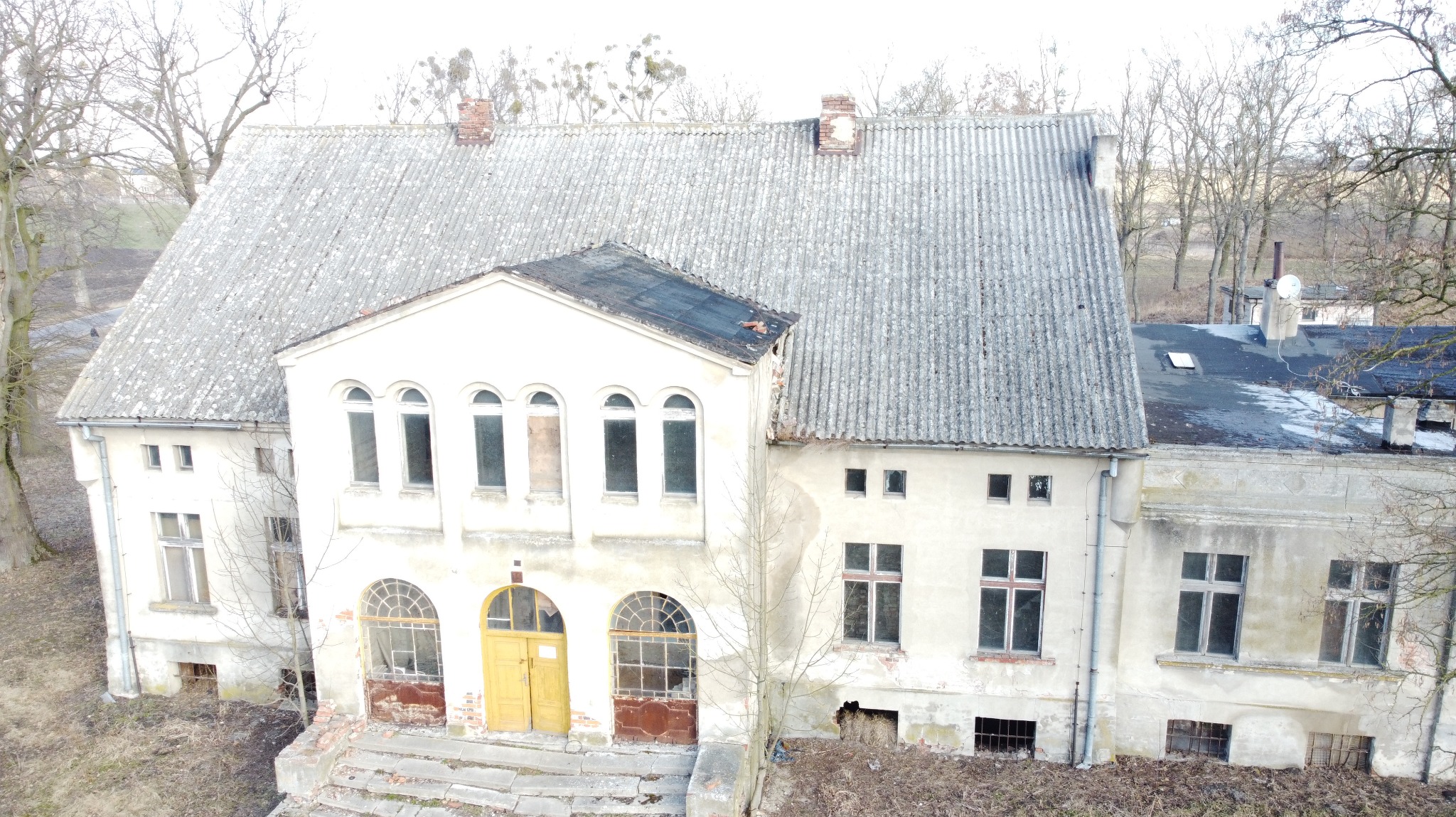 Zniszczony, dwupiętrowy budynek z szarą dachówką i żółtymi drzwiami. Widoczne uszkodzenia elewacji i dachu. Otoczenie zaniedbane, z drzewami bez liści.