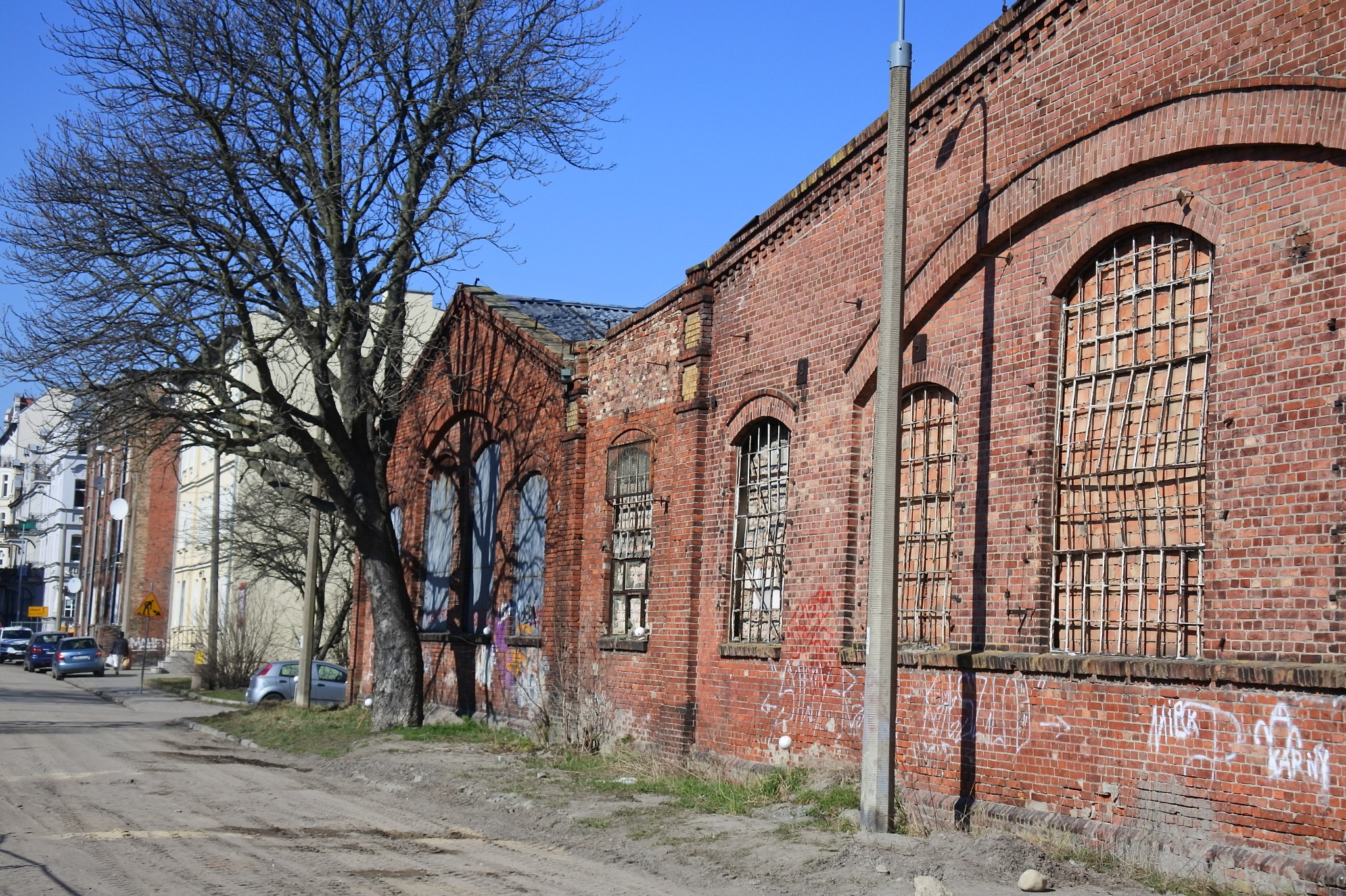 Ceglany budynek z łukowymi oknami i śladami graffiti, widoczny z ulicy z drzewem bez liści. Budynek wymaga renowacji, co sugerują uszkodzenia i brakujące fragmenty cegieł.