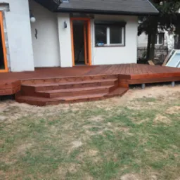 Nowo wybudowany, brązowy taras z drewna z trzema schodkami, prowadzący do domu z oknami i drzwiami w pomarańczowych ramach, na trawiastym terenie.