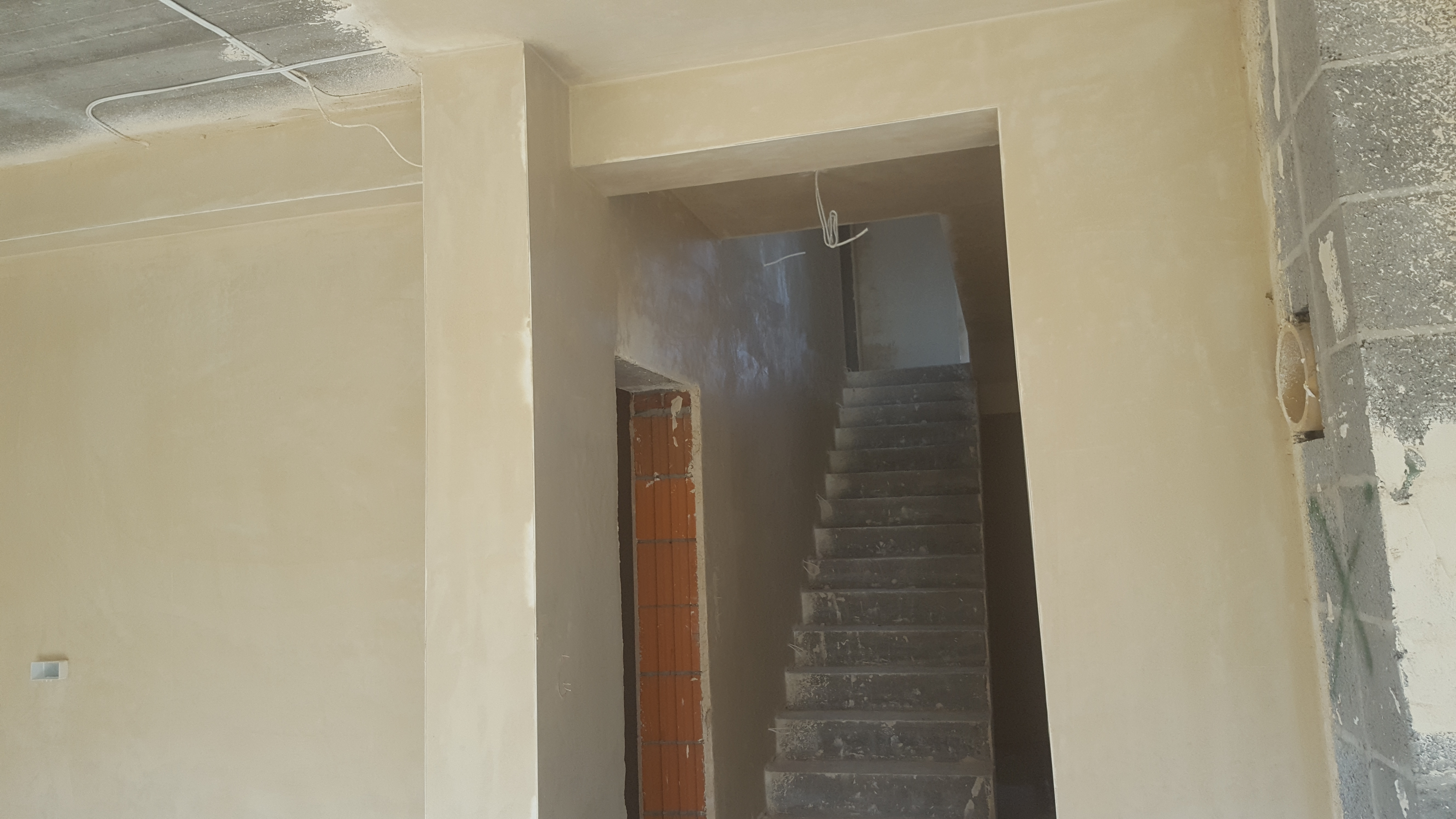 Wnętrze w trakcie prac wykończeniowych: ściany pokryte świeżym, jasnym tynkiem maszynowym, widoczne przewody elektryczne na suficie oraz surowe schody w głębi pomieszczenia.