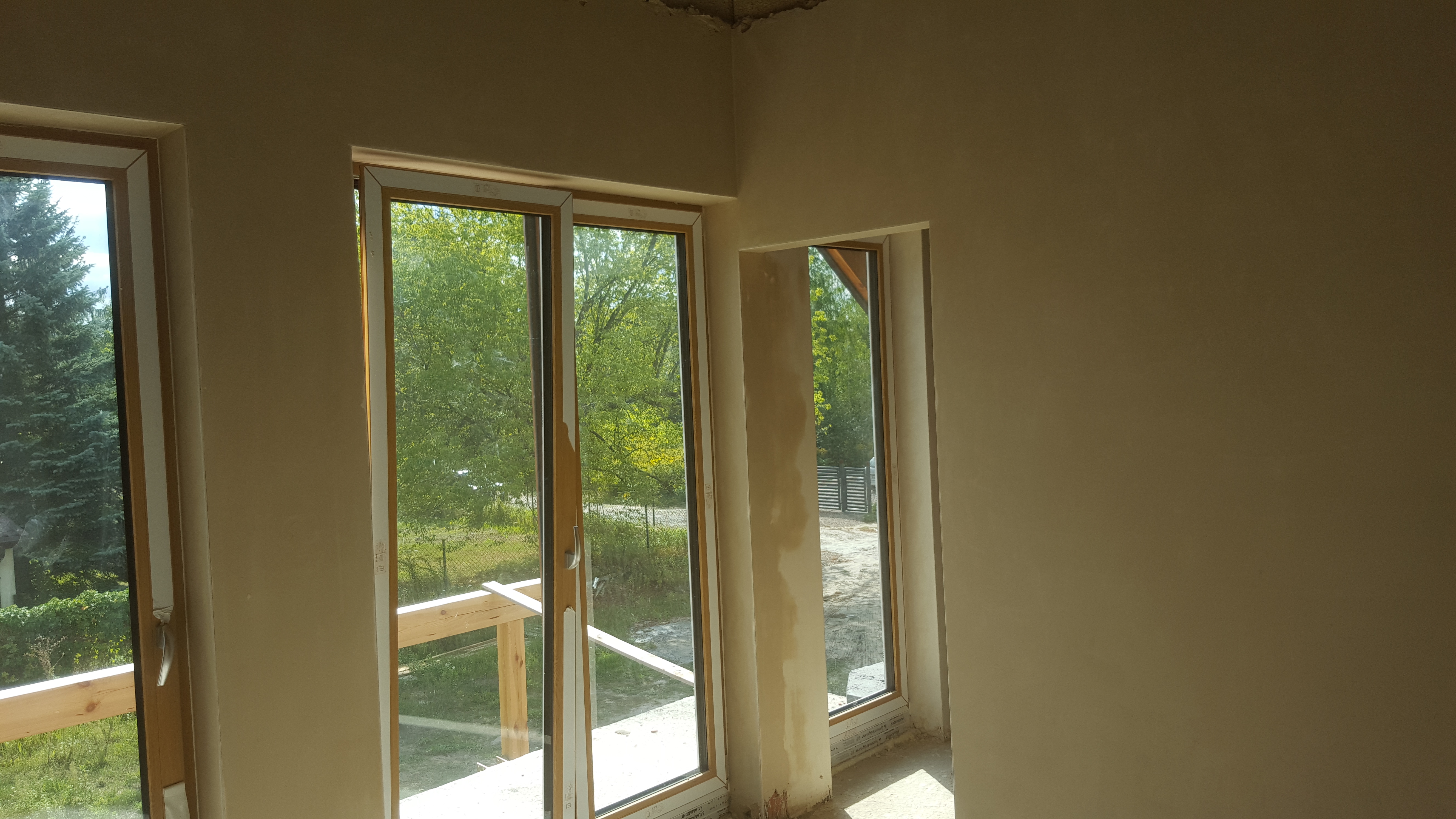 Wnętrze domu w stanie deweloperskim po tynkowaniu maszynowym, widoczne trzy okna z widokiem na zieleń i fragment surowego sufitu.