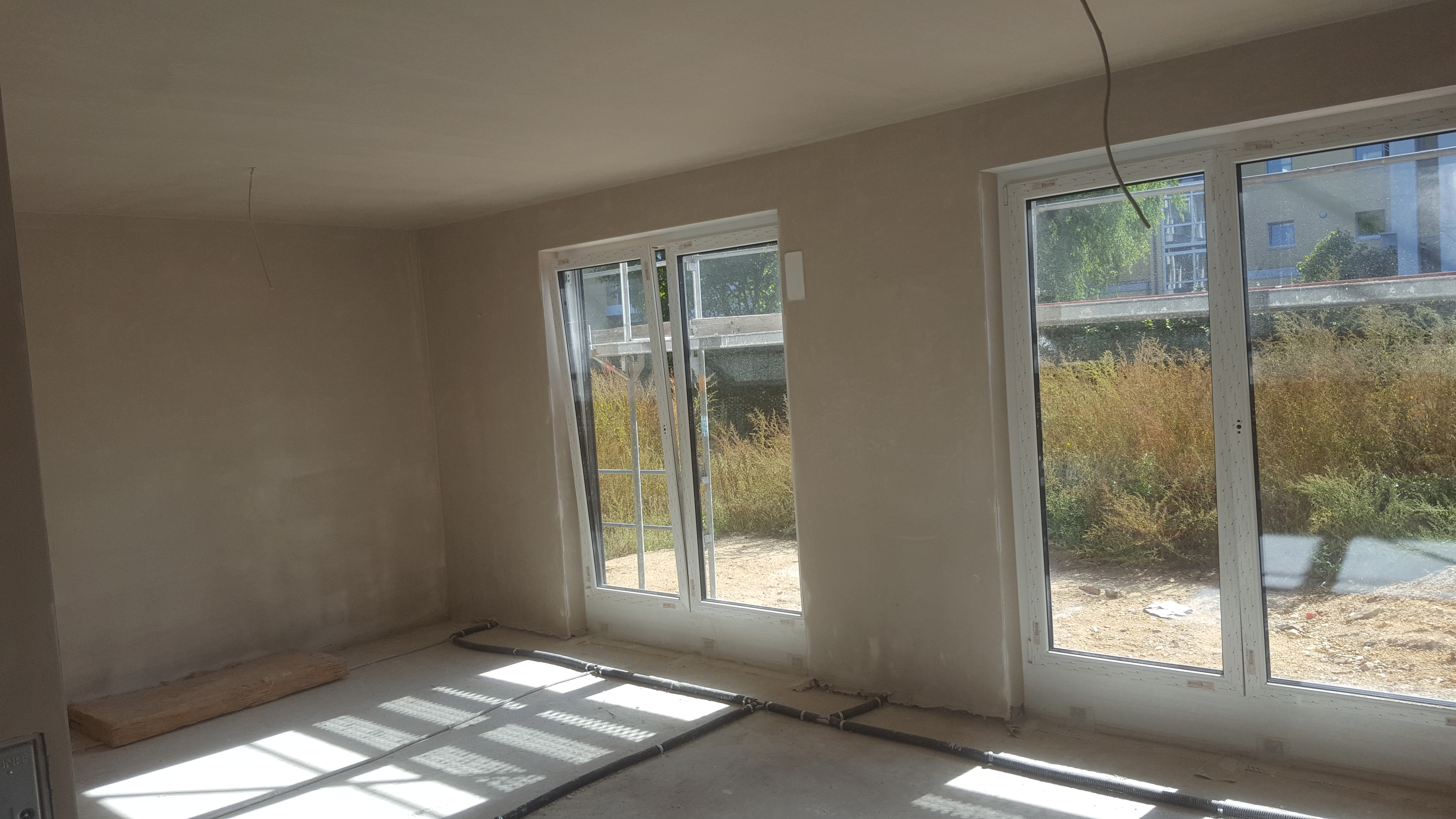 Wnętrze w stanie surowym po nałożeniu tynków maszynowych, widoczne instalacje podłogowe, dwa okna z widokiem na zewnątrz, ściany w jasnym kolorze.