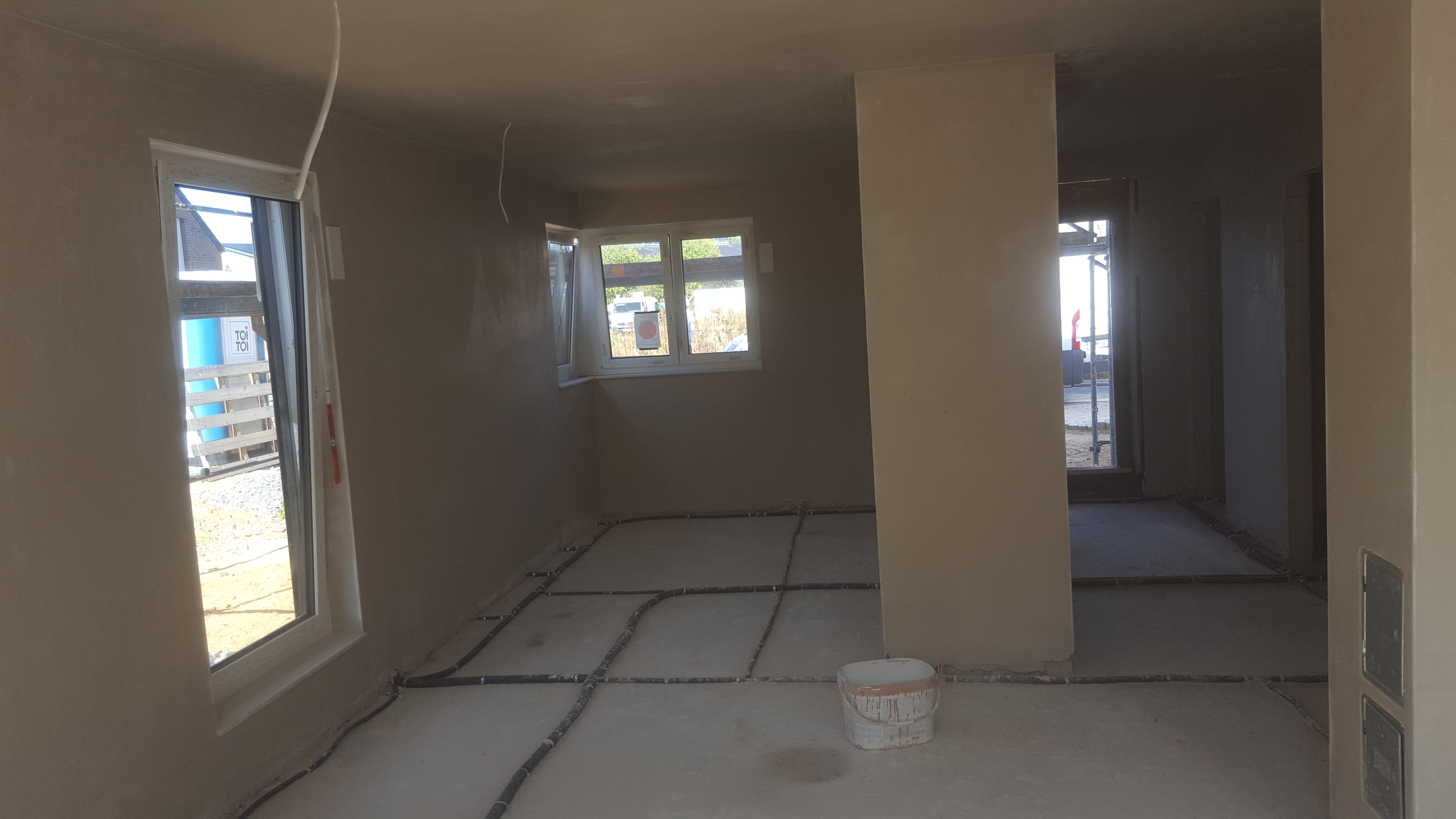 Wnętrze domu w stanie deweloperskim po nałożeniu tynków, widoczne instalacje podłogowe i wiadro po farbie.