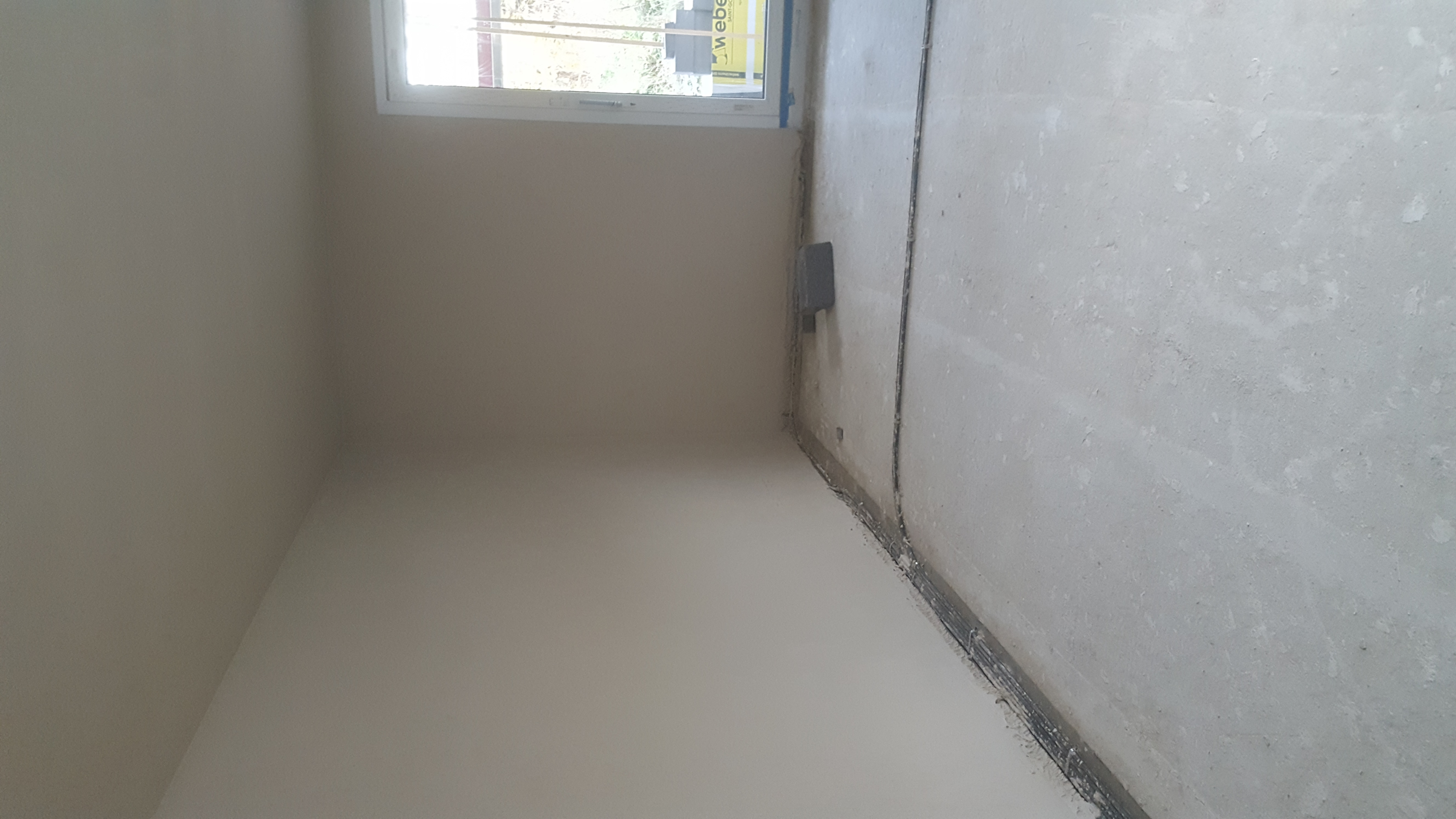 Surowe wnętrze w trakcie prac wykończeniowych, widoczna ściana po nałożeniu gładzi, surowa podłoga i wystające przewody elektryczne wzdłuż ściany, okno z widokiem na zieleń na zewnątrz.