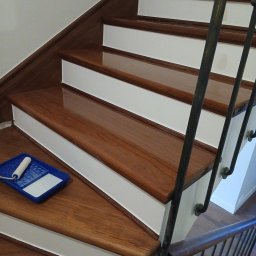 Cyklinowanie - Odnowione drewniane schody z białymi podstopnicami. Na jednym ze stopni kuweta z wałkiem malarskim. Metalowa balustrada. Połyskliwa powierzchnia drewna.