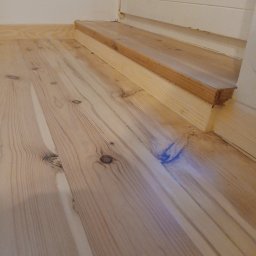 Cyklinowanie - Jasne, surowe deski podłogowe z widocznymi słojami, fragment schodka i listwy przypodłogowej. Przygotowanie do malowania lub lakierowania.