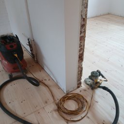 Cyklinowanie - Szlifowanie drewnianej podłogi w jasnym pomieszczeniu. Widoczna szlifierka, odkurzacz przemysłowy i zwinięty kabel. Fragment ściany z odsłoniętą cegłą.