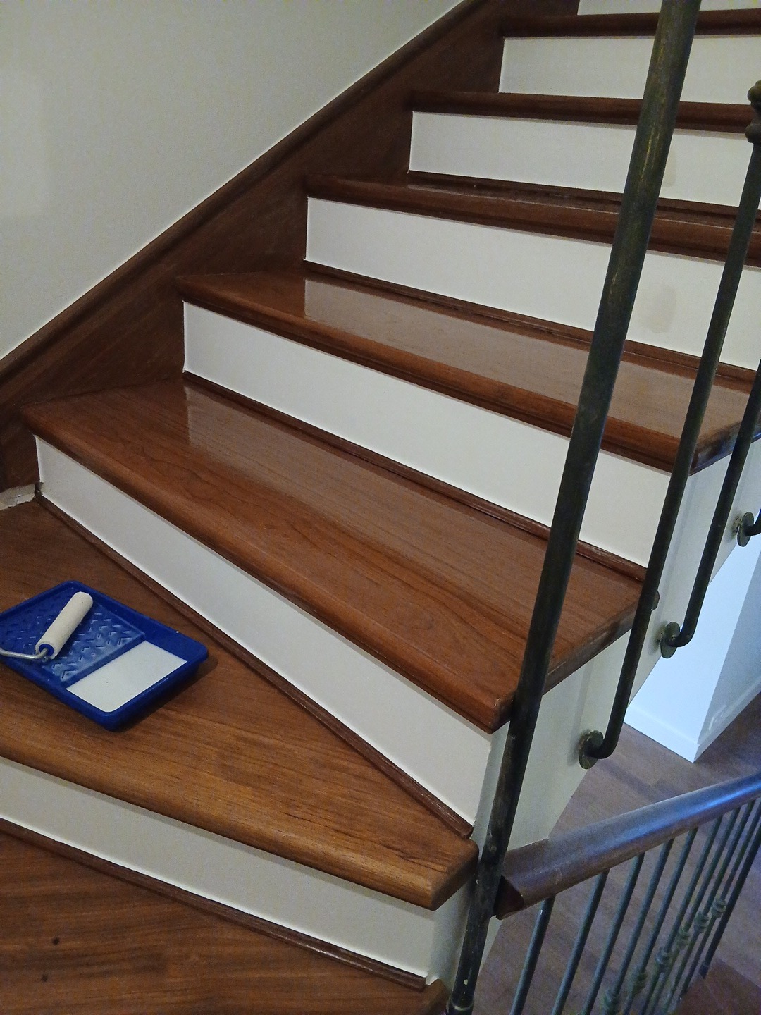 Odnowione drewniane schody z białymi podstopnicami. Na jednym ze stopni kuweta z wałkiem malarskim. Metalowa balustrada. Połyskliwa powierzchnia drewna.