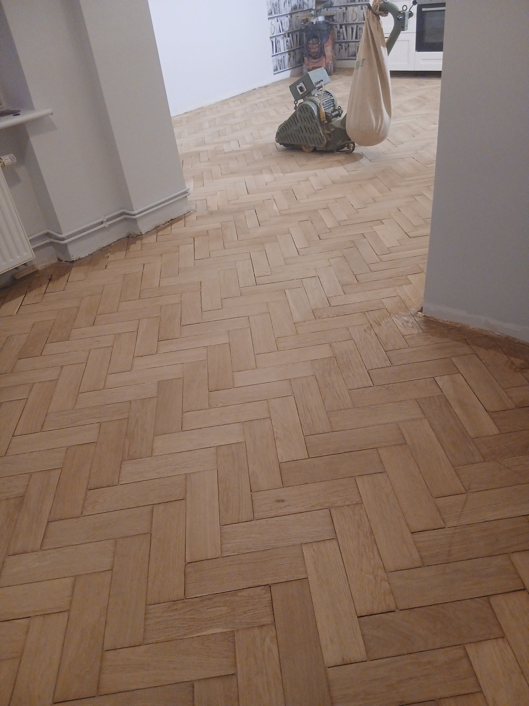 Parkiet ułożony w jodełkę po cyklinowaniu, widoczna maszyna do cyklinowania z workiem na pył. Świeżo wycyklinowana podłoga w jasnym odcieniu drewna.