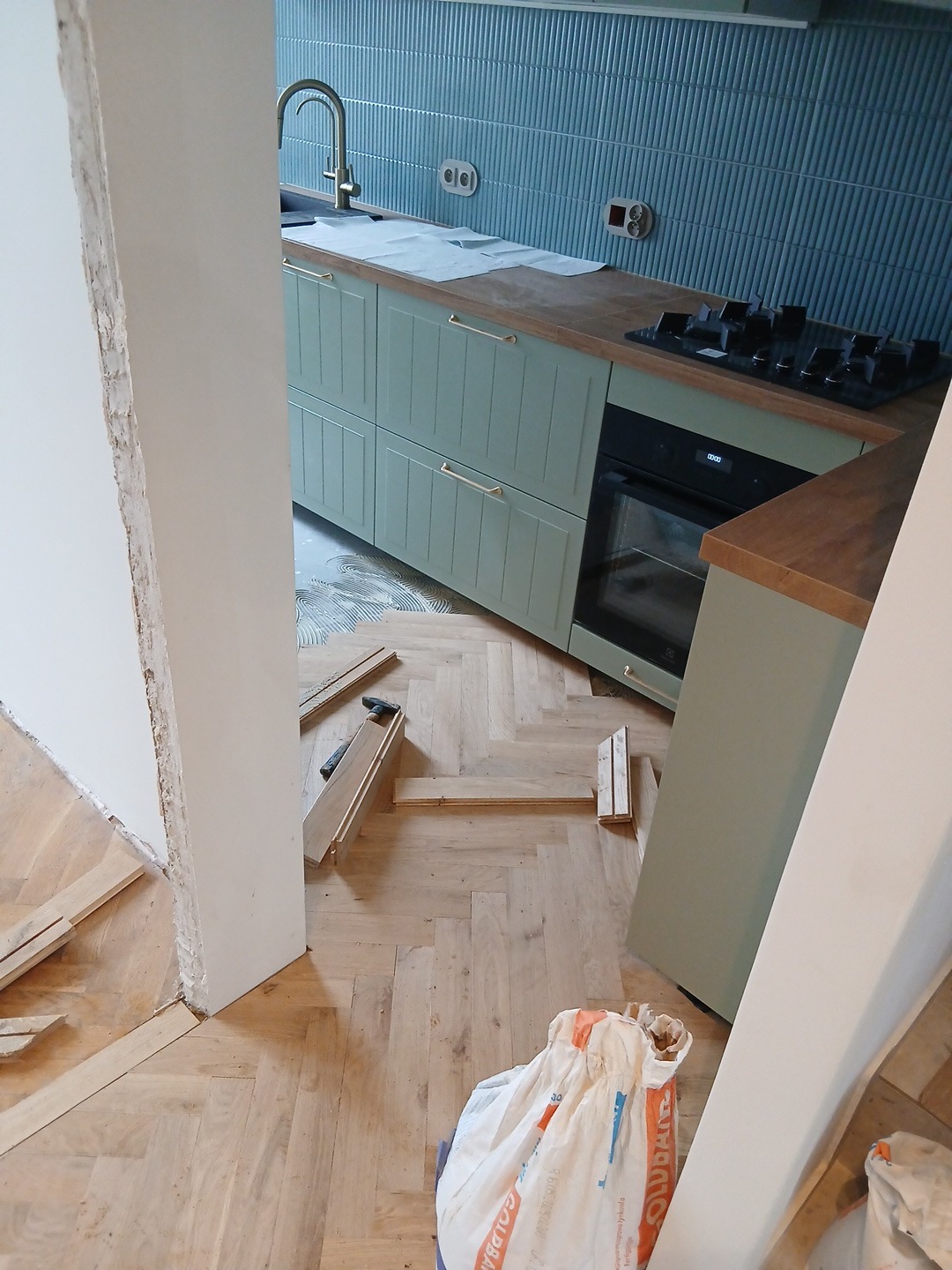 Remont kuchni: montaż parkietu w jodełkę, widoczne fragmenty zabudowy kuchennej w odcieniach zieleni i niebieska glazura na ścianie. W tle elementy wyposażenia.