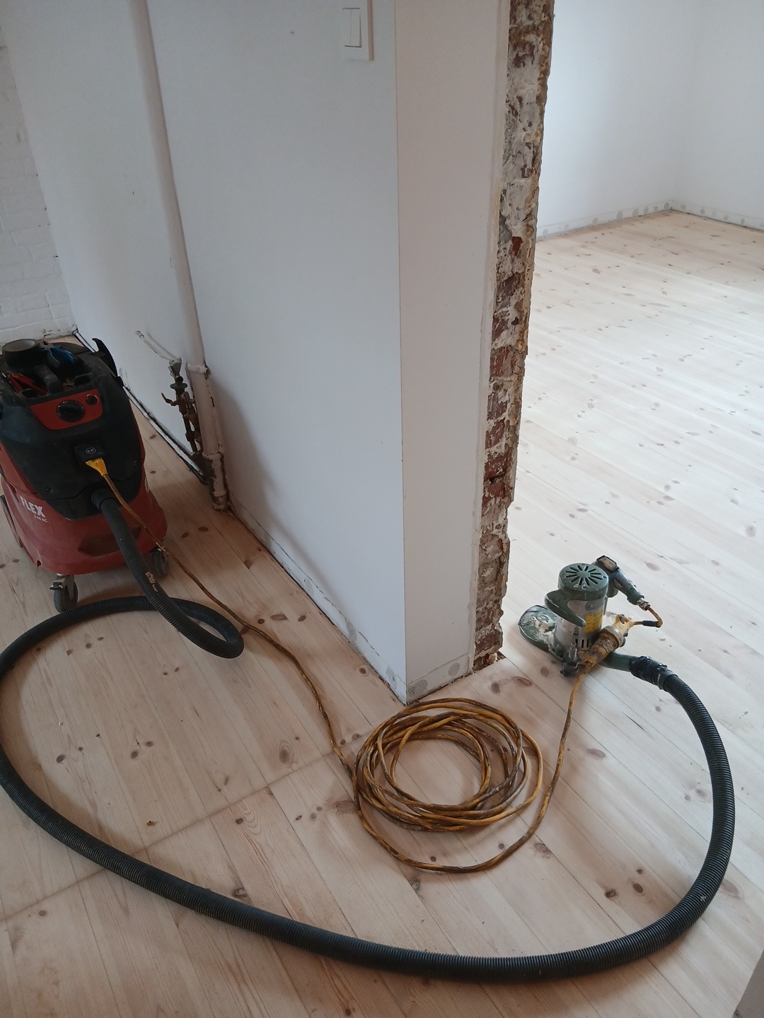 Szlifowanie drewnianej podłogi w jasnym pomieszczeniu. Widoczna szlifierka, odkurzacz przemysłowy i zwinięty kabel. Fragment ściany z odsłoniętą cegłą.