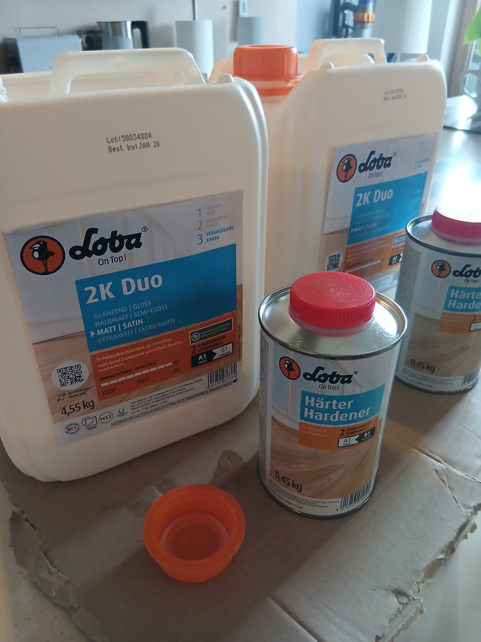 Zestaw produktów Loba: 2K Duo lakier do parkietu (4.55kg) i Härter Hardener (0.45kg). Produkty do wykończenia podłóg drewnianych, widoczne etykiety z opisami i datami ważności.