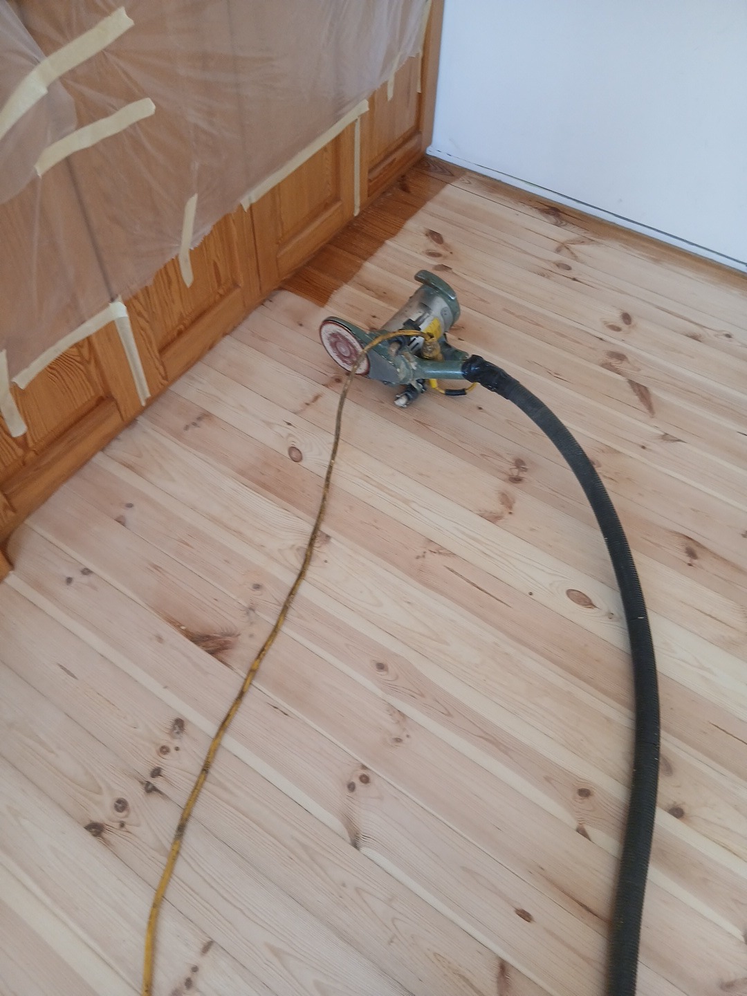 Szlifowanie drewnianej podłogi przy użyciu szlifierki oscylacyjnej. Część podłogi już polakierowana, widoczne zabezpieczenie folią i taśmą malarską przy listwach.