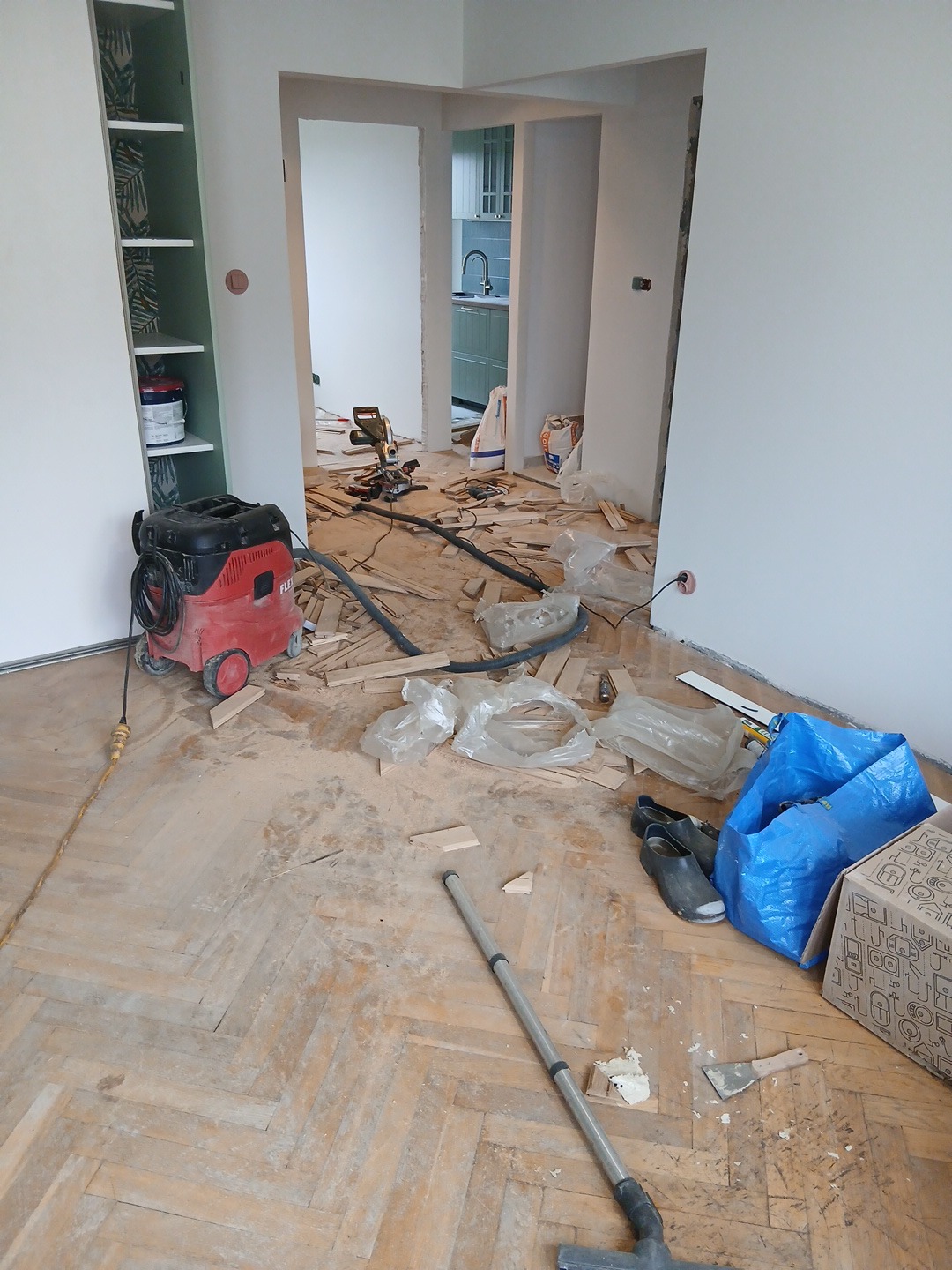 Remont mieszkania: podłoga w jodełkę po demontażu, widoczny odkurzacz przemysłowy, narzędzia i resztki materiałów. W tle odnowiona kuchnia.