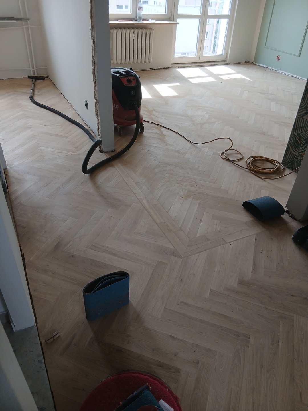 Parkiet ułożony w jodełkę po cyklinowaniu, z widocznym odkurzaczem przemysłowym i materiałami ściernymi. Jasne wnętrze z oknem i grzejnikiem.