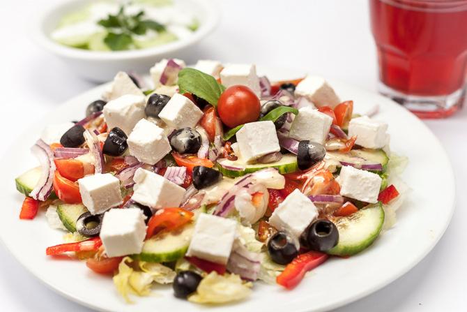 Sałatka grecka z kostkami sera feta, czarnymi oliwkami, pomidorami, ogórkiem, czerwoną cebulą i papryką na białym talerzu, z miseczką sosu i szklanką czerwonego napoju w tle.