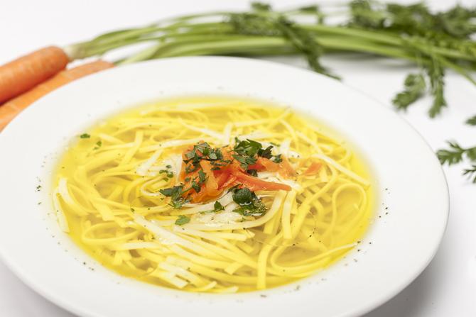 Talerz rosołu z makaronem, marchewką i natką pietruszki, z boku widoczne marchewki z zielonymi liśćmi.