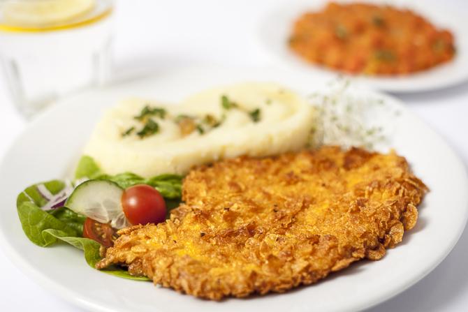 Panierowany kotlet z puree ziemniaczanym posypanym ziołami i sałatką ze świeżych warzyw na białym talerzu, w tle drugi talerz z pomidorowym daniem.