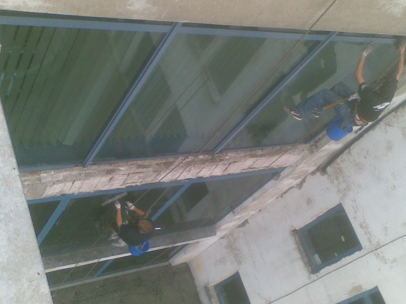 Dwóch alpinistów na linach myje szklany dach nad patio, widoczny wiadro i ściągaczka.