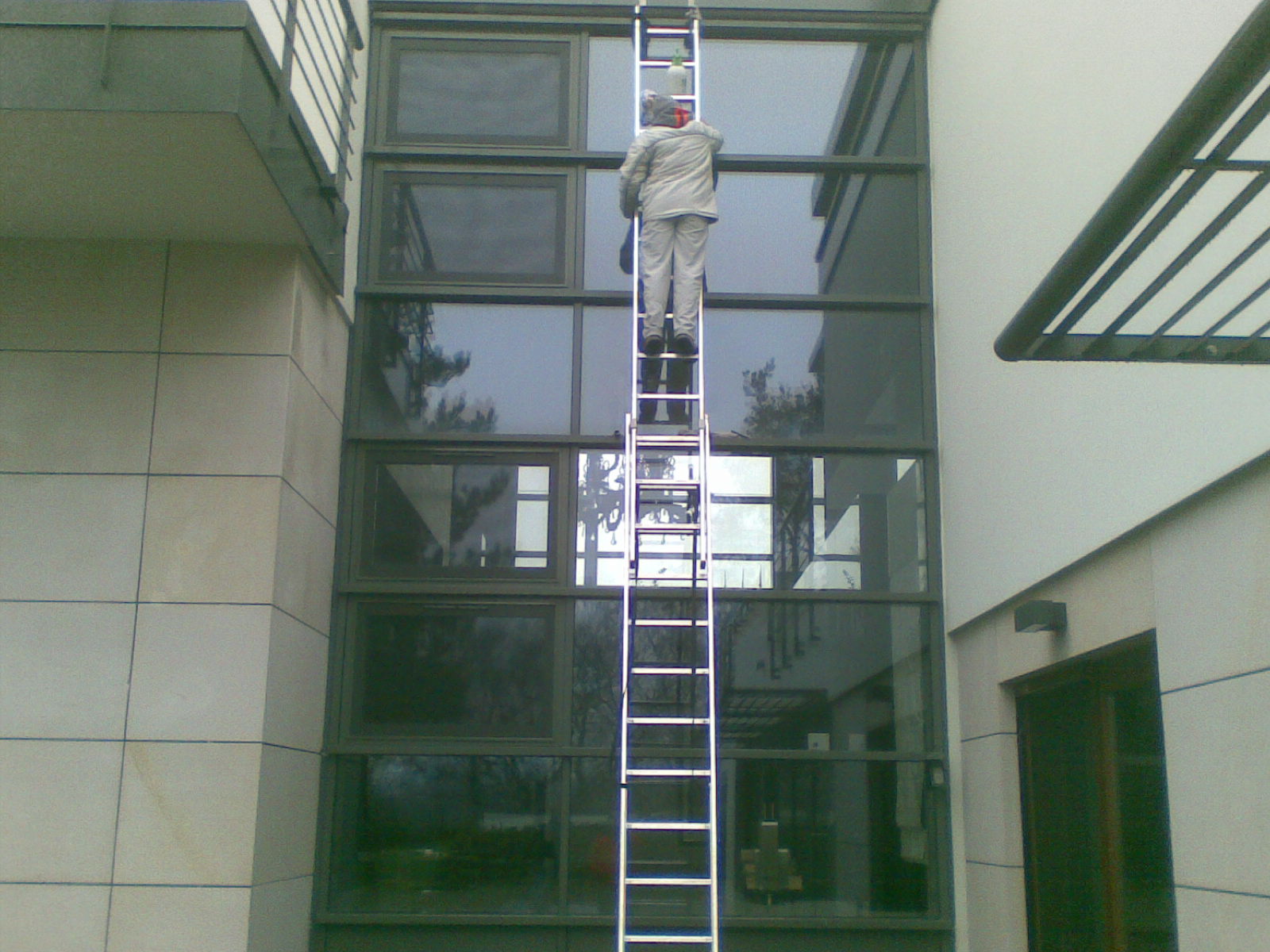 Pracownik w kombinezonie na wysokiej aluminiowej drabinie, myjący okna w nowoczesnym biurowcu z dużymi przeszkleniami, widok z dołu.