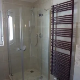 Kabina prysznicowa narożna z przezroczystego szkła, zamontowana w łazience wyłożonej beżowymi płytkami, obok grzejnik drabinkowy w kolorze ciemnej czekolady.