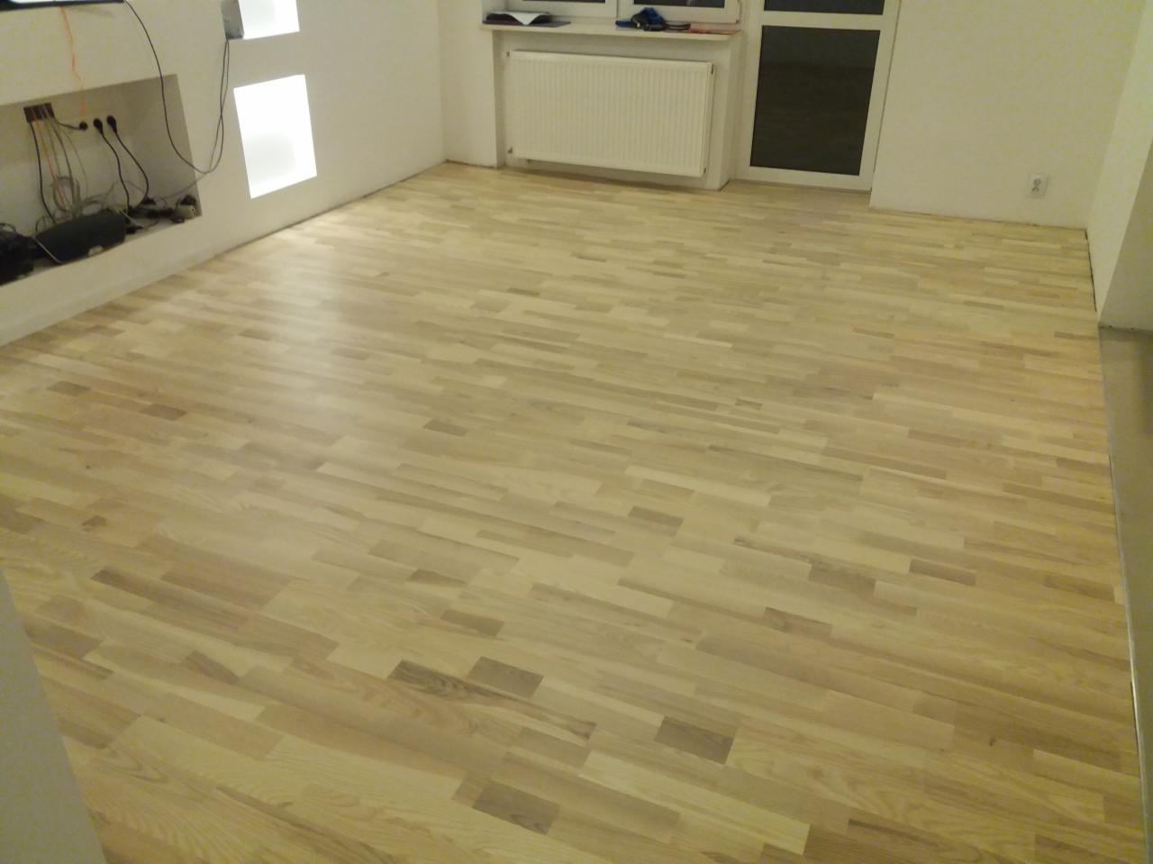 Jasna, nowa podłoga z parkietu ułożonego w jodełkę francuską w pustym pokoju z białymi ścianami i grzejnikiem pod oknem.