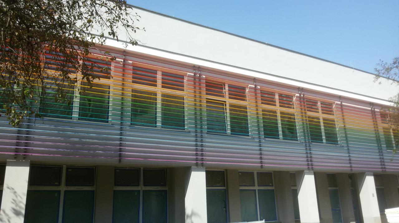 Fasada budynku z poziomymi, kolorowymi żaluzjami okiennymi w barwach tęczy, widoczne fragmenty drzew i błękitne niebo.