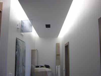 Korytarz z białym sufitem podwieszanym z oświetleniem LED po bokach i otworem na lampę, zabezpieczona folią rama drzwiowa i materiały budowlane w tle.