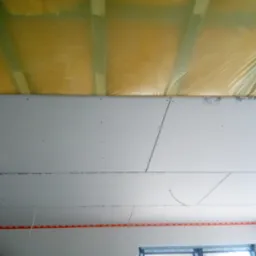 Montaż sufitu podwieszanego z płyt kartonowo-gipsowych, widoczna izolacja z folii i wełny mineralnej oraz taśma miernicza na ścianie.