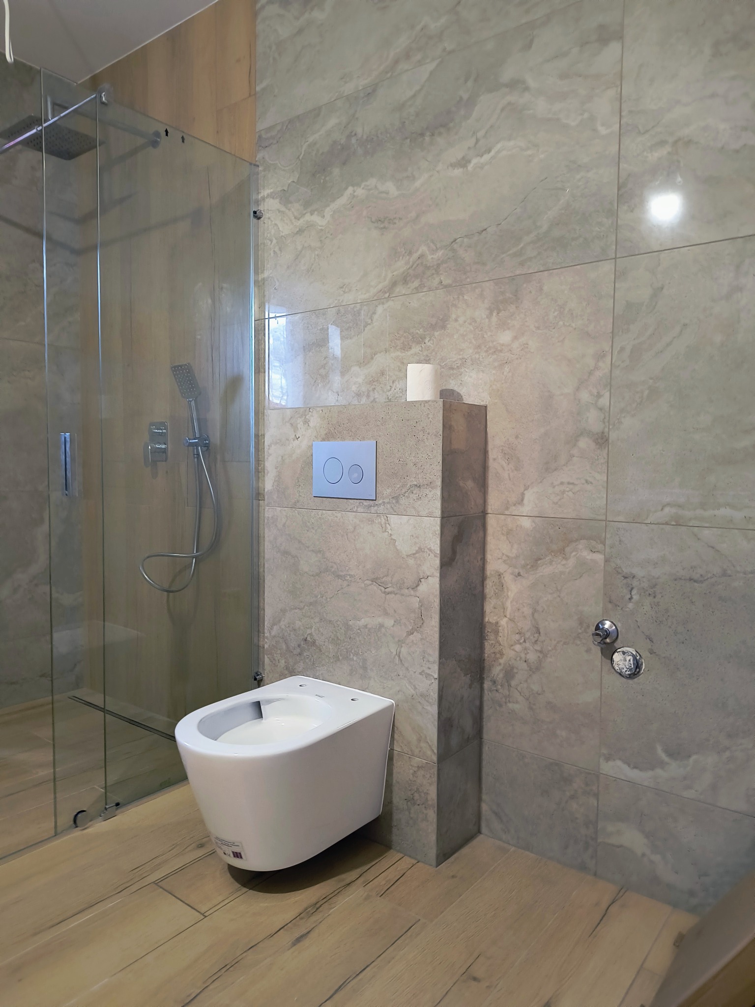 Nowoczesna łazienka z kabiną prysznicową ze szkła, podwieszanym bidetem, marmurowymi płytkami na ścianach i drewnopodobną podłogą, z rolką papieru toaletowego na obudowie stelaża.