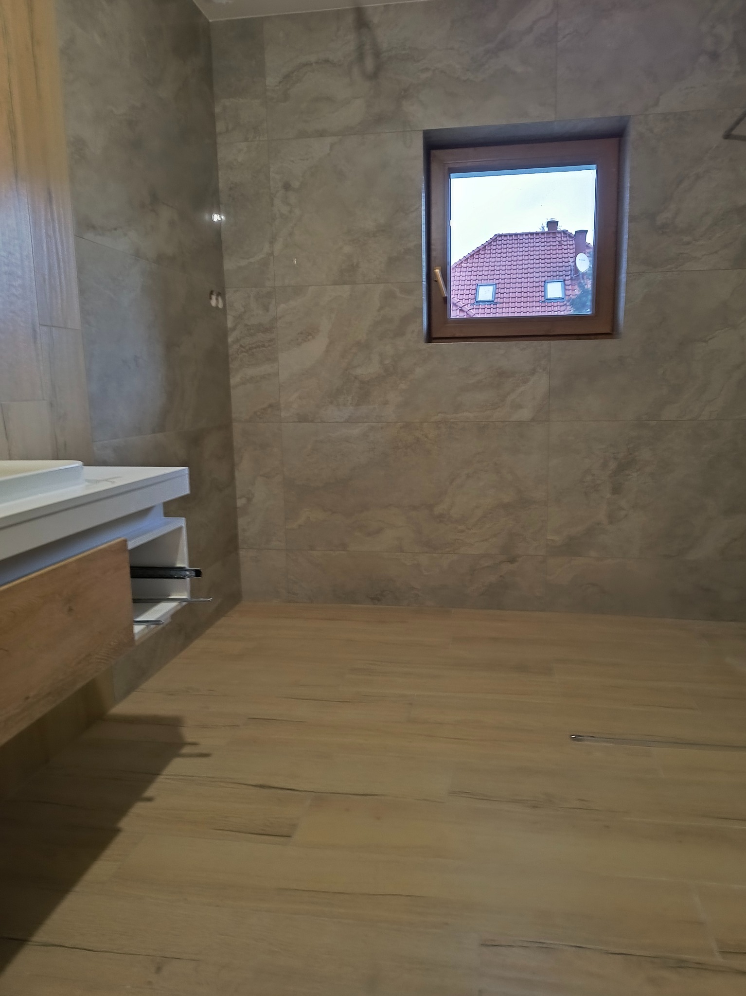 Wykończona łazienka z beżowymi płytkami imitującymi kamień na ścianach i podłodze, wisząca szafka pod umywalkę z drewnianym frontem i widokiem na dom z czerwonym dachem przez okno.