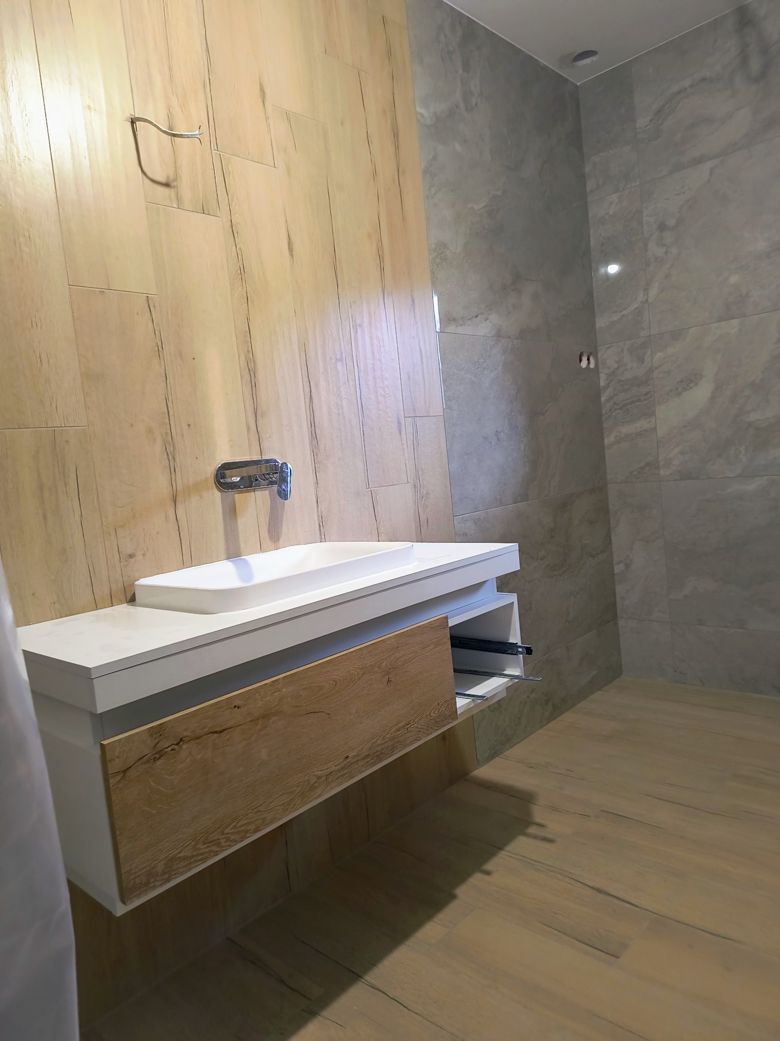 Nowoczesna łazienka z drewnopodobnymi płytkami na ścianie i podłodze, białą umywalką nablatową z szafką z drewnianym frontem i baterią chromowaną, szare płytki imitujące marmur w strefie prysznicowej.
