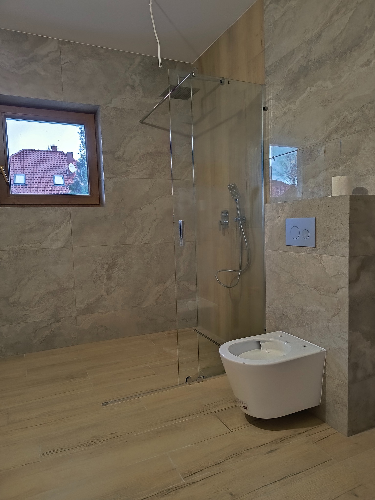 Nowoczesna łazienka z kabiną prysznicową typu walk-in, podwieszanym bidetem, płytkami imitującymi marmur i oknem z widokiem na czerwoną dachówkę.