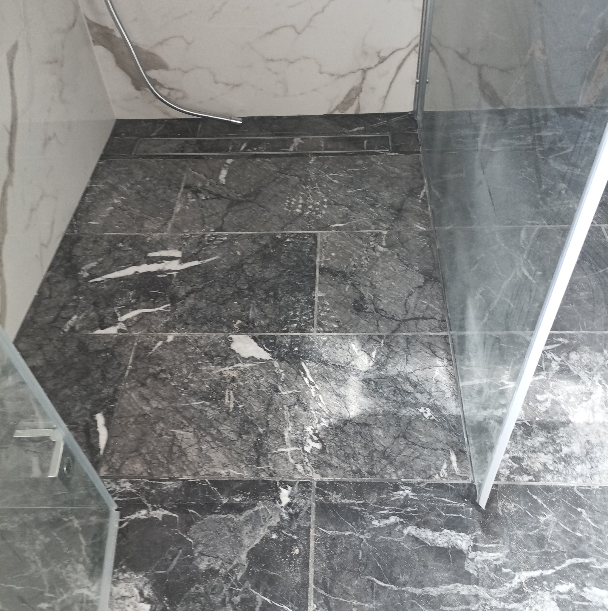 Wykończona podłoga w łazience z ciemnoszarych płytek imitujących marmur, z widocznym odpływem liniowym i fragmentem kabiny prysznicowej z przezroczystego szkła.