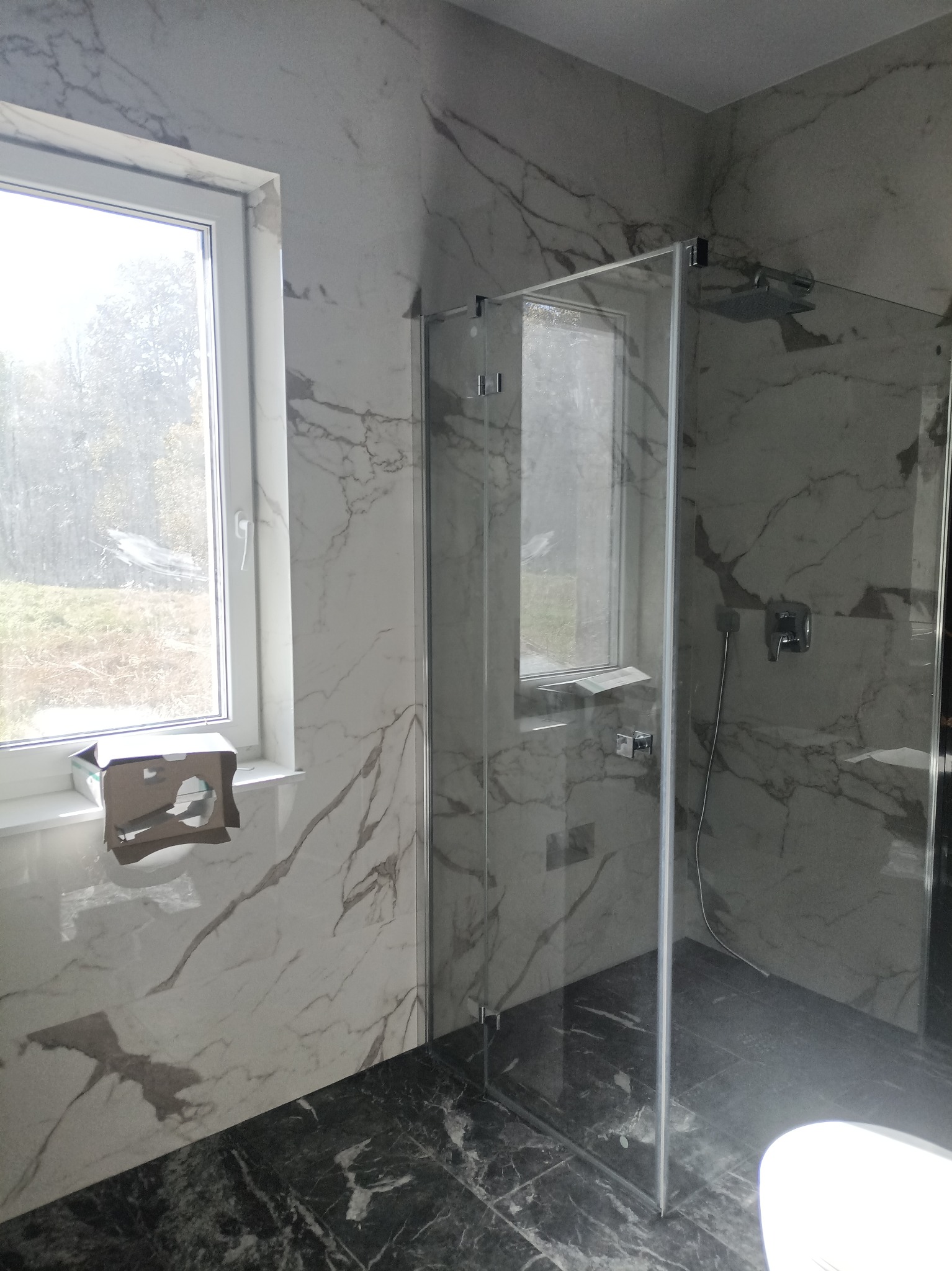 Nowoczesna kabina prysznicowa ze szklanymi ściankami i deszczownicą, marmurowe płytki na ścianach i podłodze, widok na okno z kartonem na parapecie.