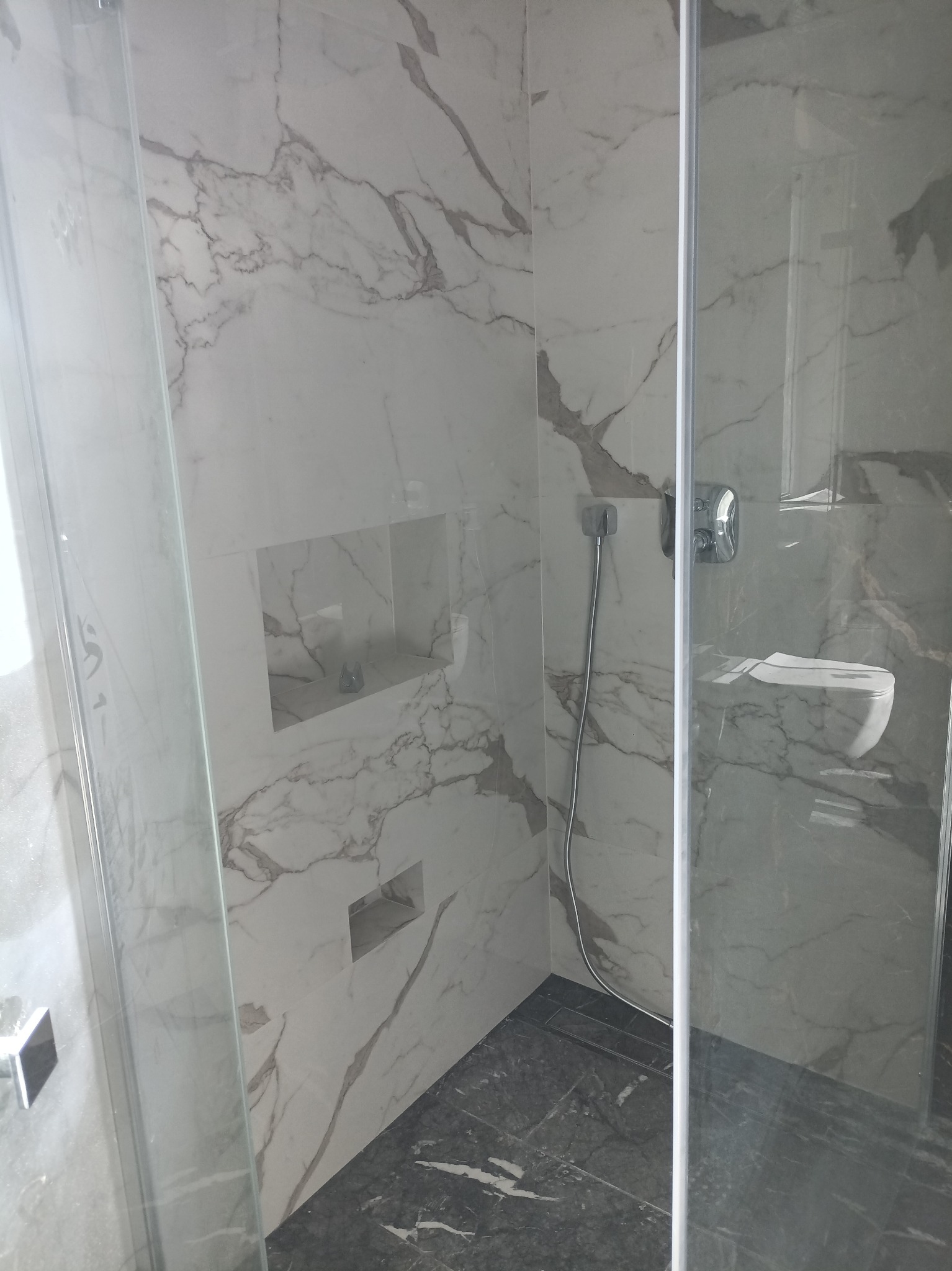 Wykończona łazienka z kabiną prysznicową typu walk-in, wyłożona białymi płytkami imitującymi marmur z szarymi żyłami, z wnękami w ścianie i ciemnoszarą podłogą z odpływem liniowym.