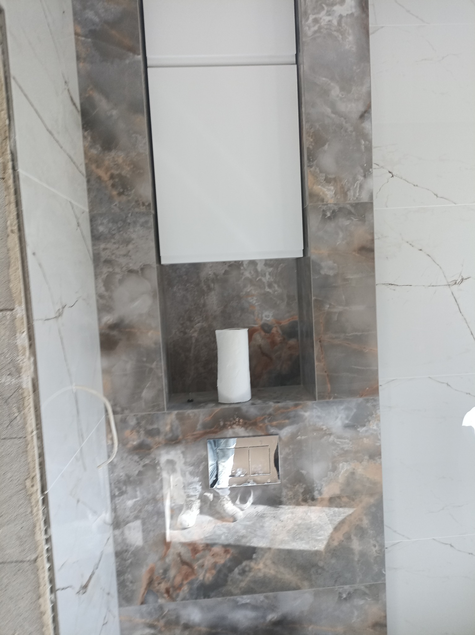Nisza w łazience wyłożona płytkami imitującymi marmur, z rolką papieru toaletowego i przyciskiem spłuczki WC. Obok widoczna ściana z białych płytek z szarymi żyłkami i fragment surowej ściany.