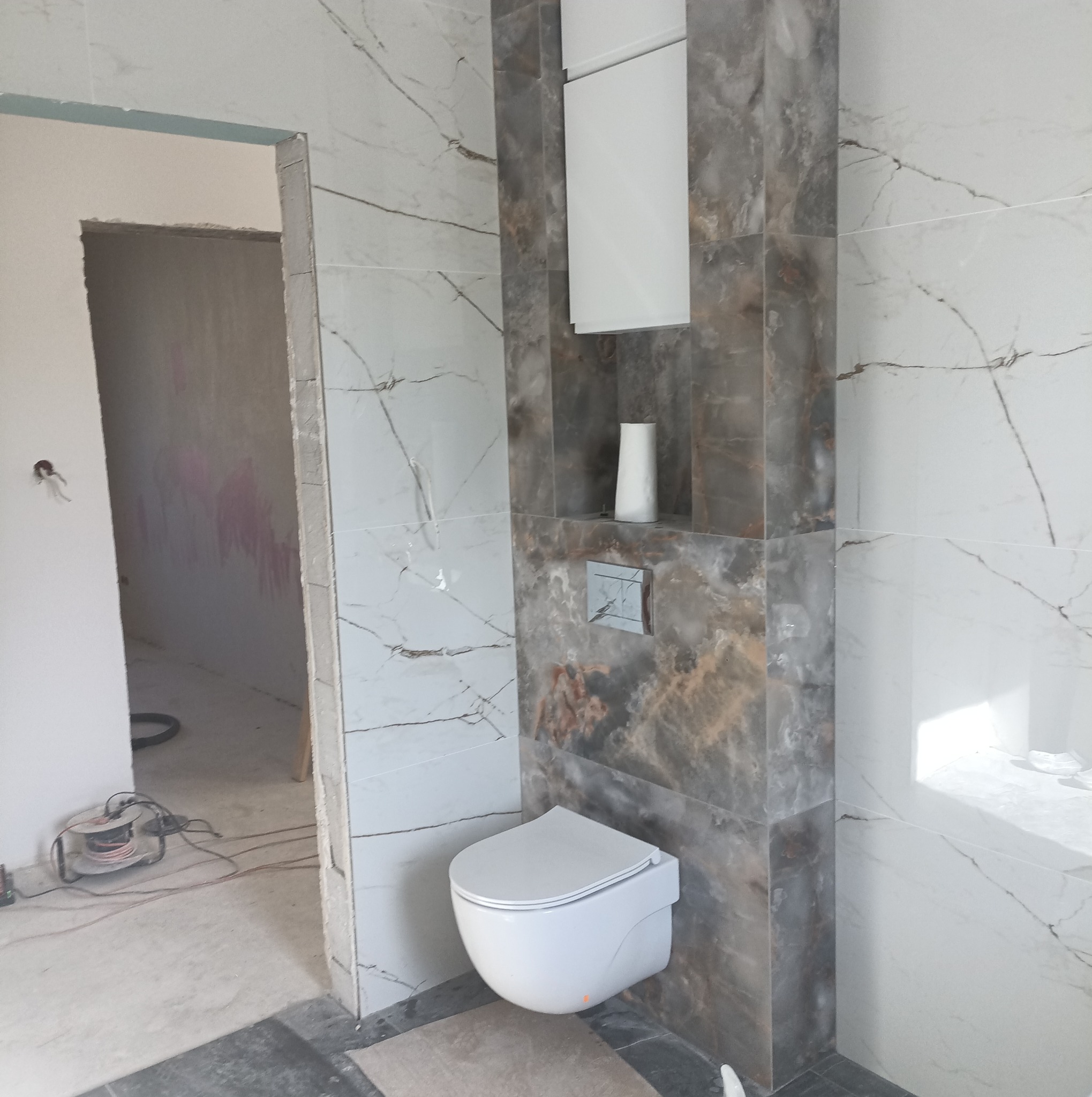 Wnętrze łazienki w trakcie prac: podwieszana toaleta, zabudowa z płytek imitujących marmur, widoczny przewód elektryczny i surowe ściany w tle.