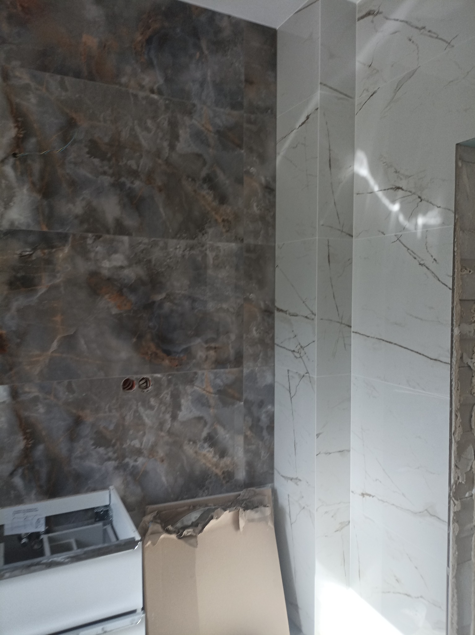 Ściana łazienki wyłożona dwoma rodzajami płytek: szare płytki imitujące marmur z widocznymi otworami na gniazdka oraz białe płytki z szarymi żyłkami, narożnik wykończony listwą. Częściowo widoczna...