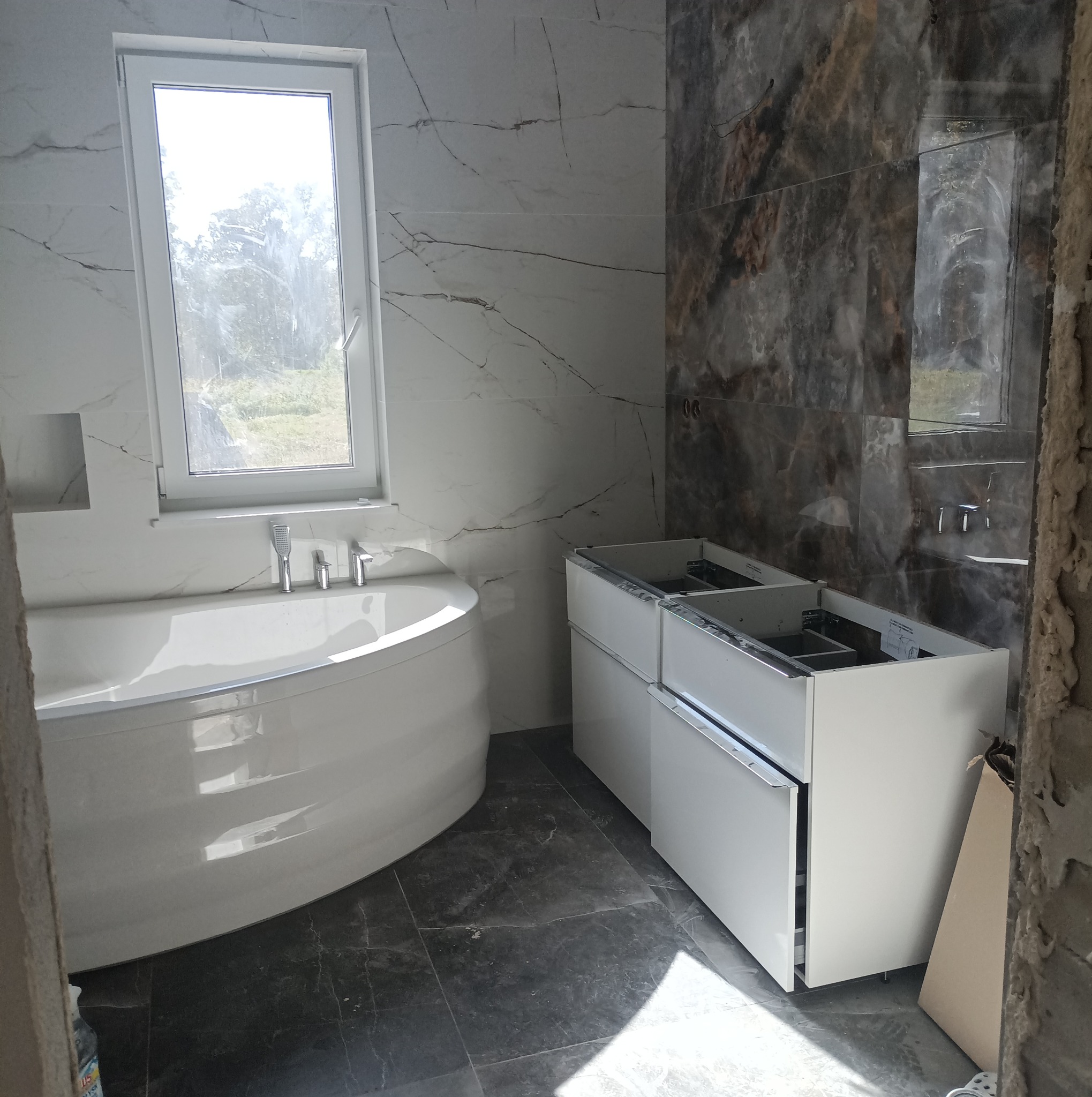 Nowoczesna łazienka w trakcie wykańczania: biała wanna pod oknem, marmurowe płytki ścienne w dwóch odcieniach oraz szafka pod umywalkę bez blatu.