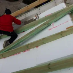 Ocieplanie dachu budynku: mężczyzna w czerwonej kurtce montuje białe płyty izolacyjne między drewnianymi belkami, używając różowej pianki montażowej.