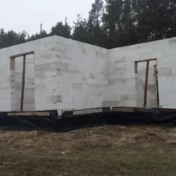 Ściany parterowego domu z białych bloczków, z widocznymi otworami okiennymi i drzwiowymi, na fundamencie z czarną izolacją, w tle drzewa.