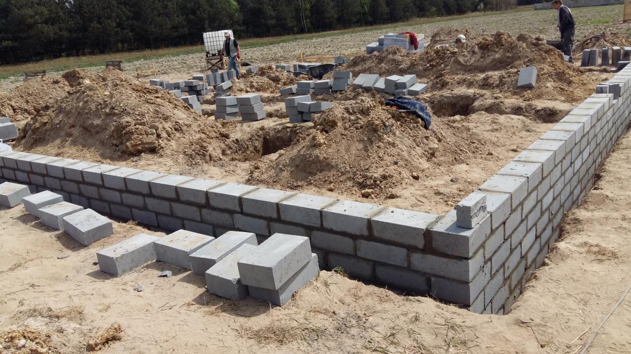Budowa fundamentów z bloczków betonowych. Częściowo wzniesione ściany fundamentowe na placu budowy z widocznymi stertami ziemi i pracownikami w tle.