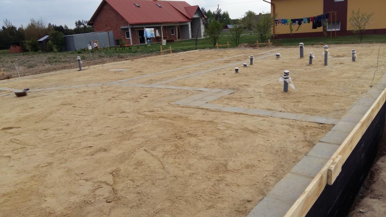 Przygotowany teren pod budowę domu z widocznymi fundamentami, izolacją i pionami kanalizacyjnymi. W tle budynki mieszkalne.