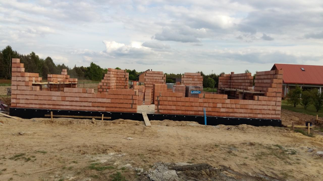 Wznoszenie ścian z czerwonej cegły na fundamencie, widoczne zbrojenia i elementy konstrukcyjne, w tle budynek z czerwonym dachem i drzewa.