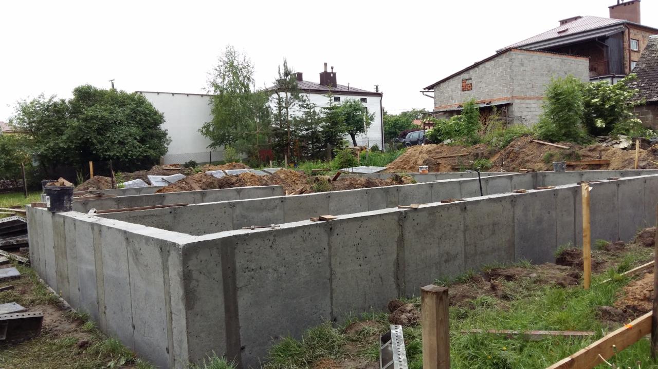 Betonowe fundamenty domu na działce z zieloną trawą, widok na budynki mieszkalne w tle. Etap budowy domu, szalunki przygotowane pod wylewkę.