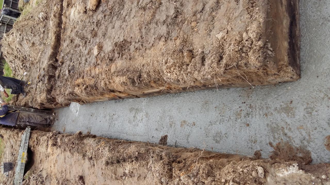 Wylany fundament w wykopie, widoczne ślady pracy sprzętu budowlanego, ziemia i świeży beton. Ujęcie z góry, dokumentujące etap budowy.